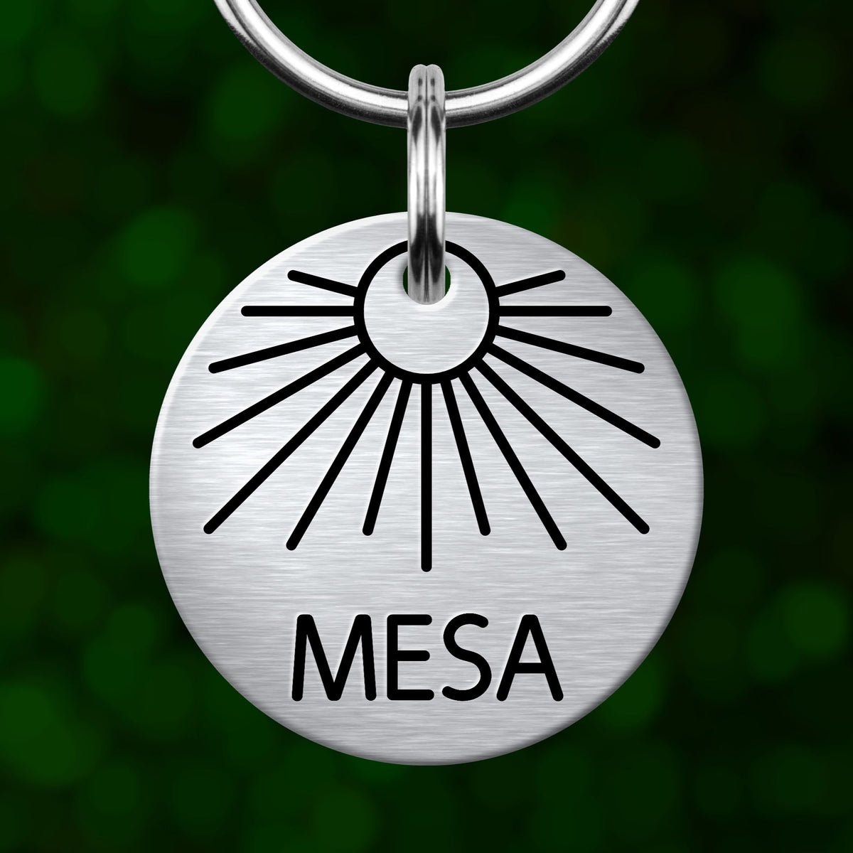 A silver-colored metal keychain with a sun-like design and the word "MESA" printed on it.