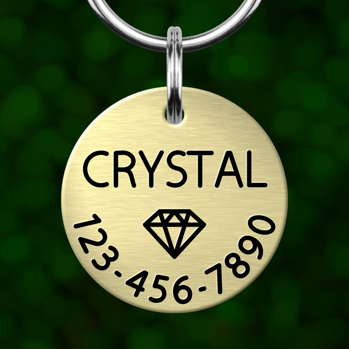 A gold-colored metal pet tag with the name "CRYSTAL" and a diamond symbol, along with the numbers "123-456-7890".