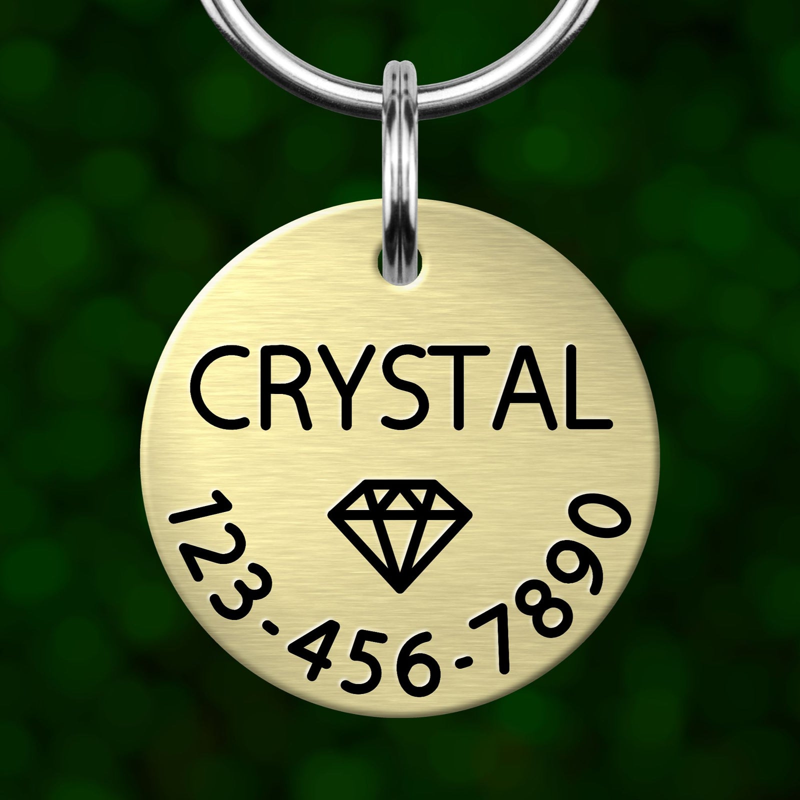 A gold-colored metal pet tag with the name "CRYSTAL" and a diamond symbol, along with the numbers "123-456-7890".