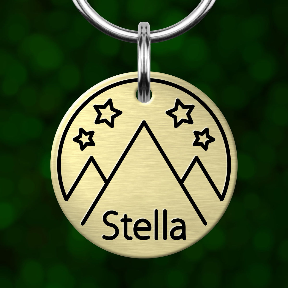 A round metal keychain tag with a mountain and star design, and the name "Stella" written on it.
