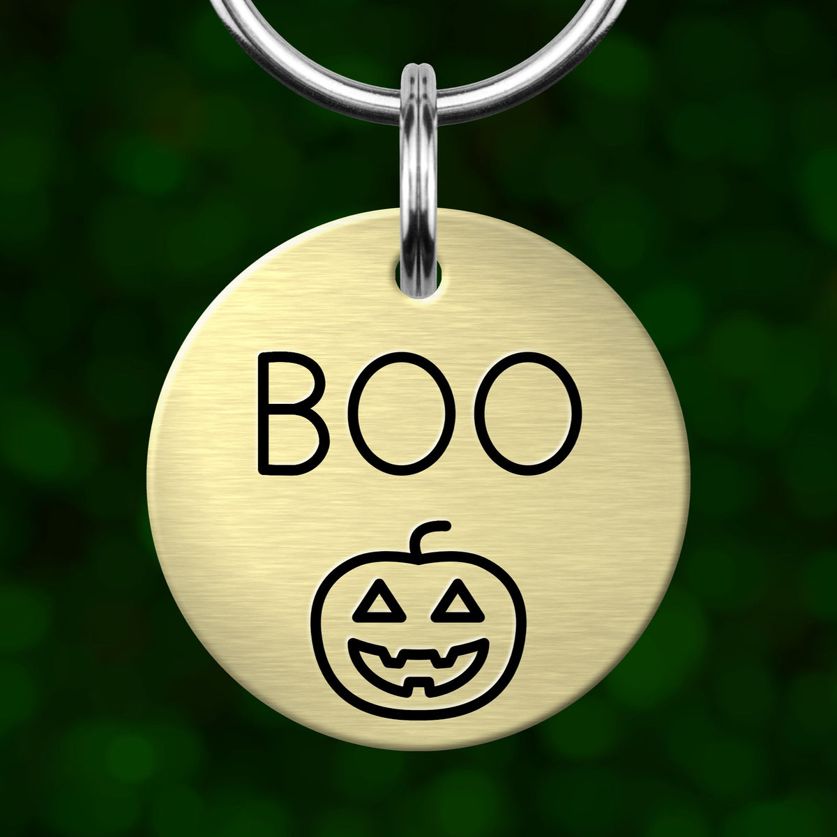 A round metal keychain tag with the word "BOO" and a pumpkin icon printed on it, set against a blurred green background.