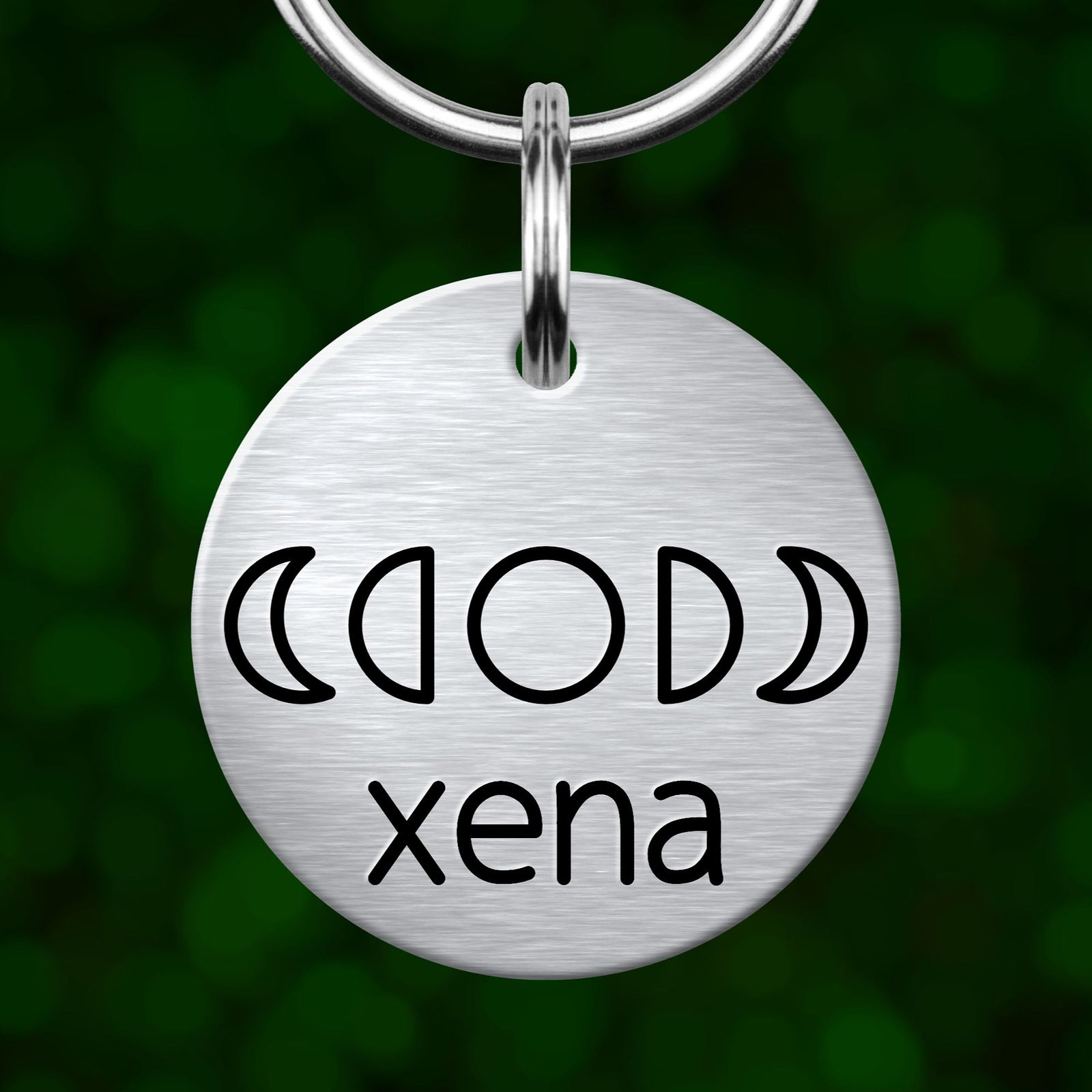 A silver metal keychain tag with the name 'Xena' written on it, featuring a design of the moon phases.