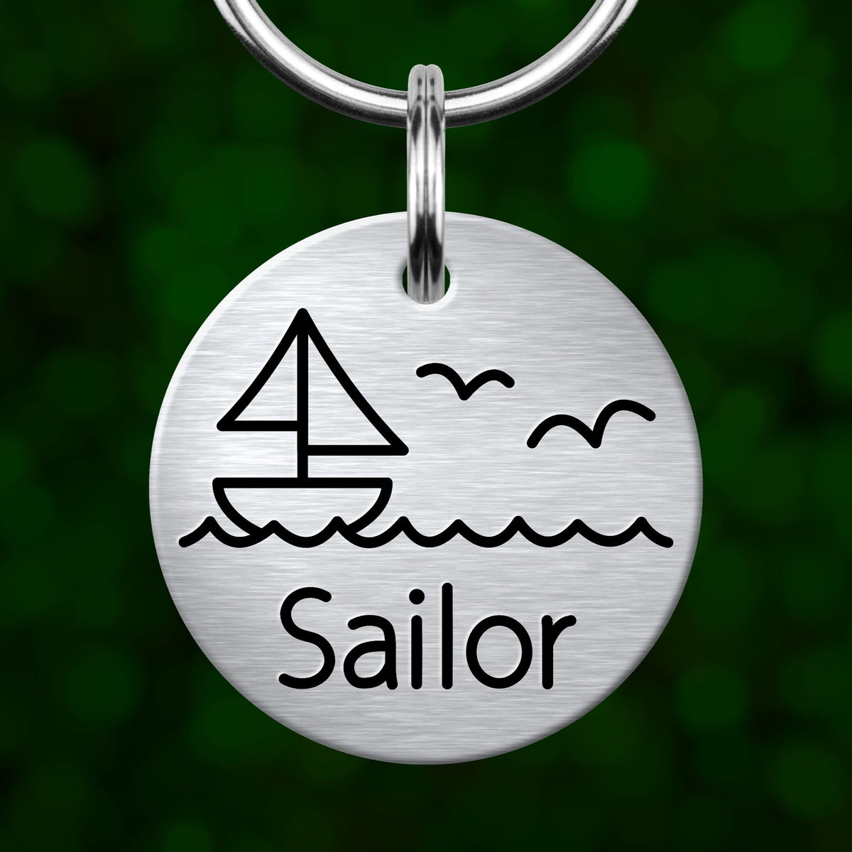A round metal keychain tag with a graphic of a sailboat and waves, along with the word "Sailor" written on it.