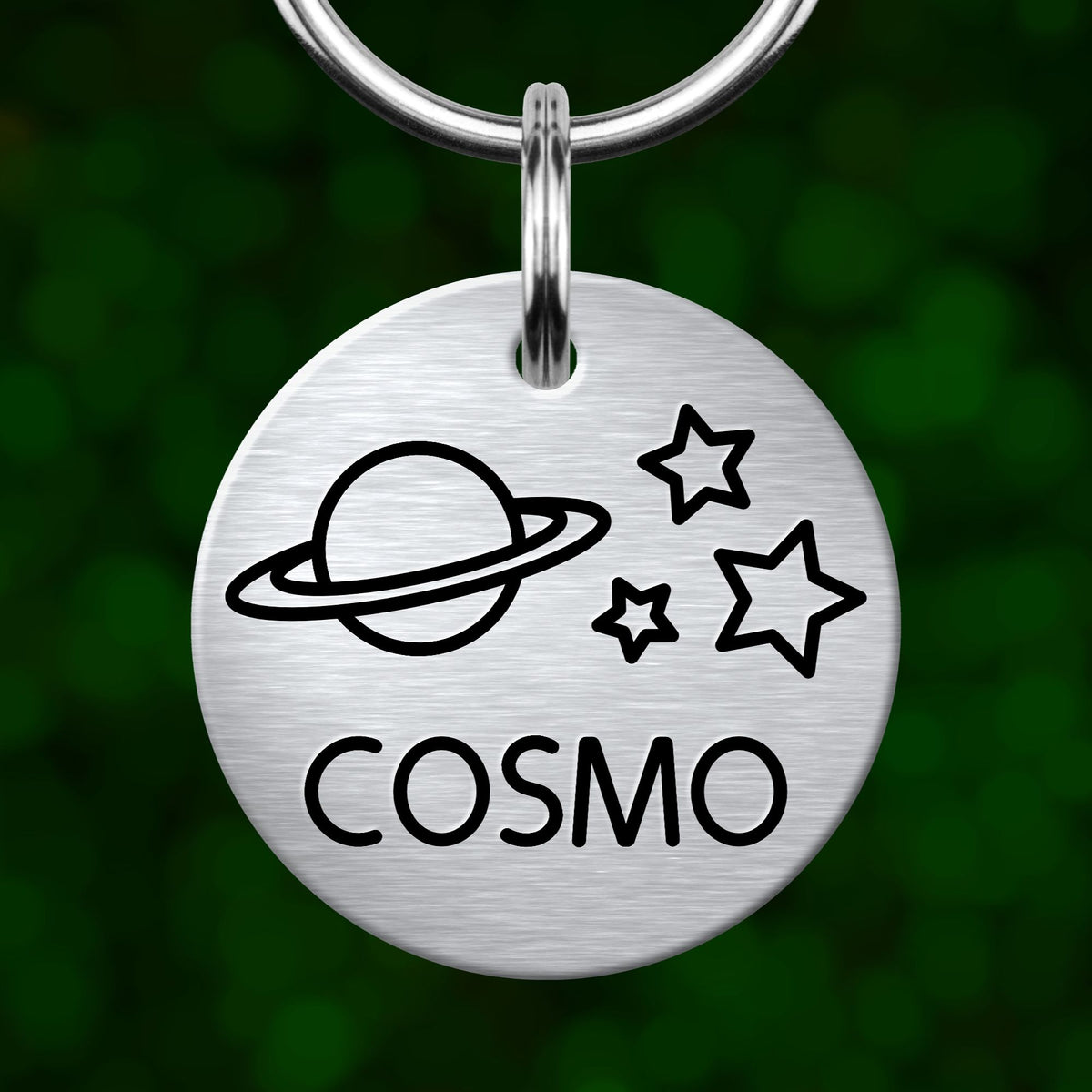 A silver circular pet tag with a space-themed design, including a planet, stars, and the word "COSMO" printed on it.