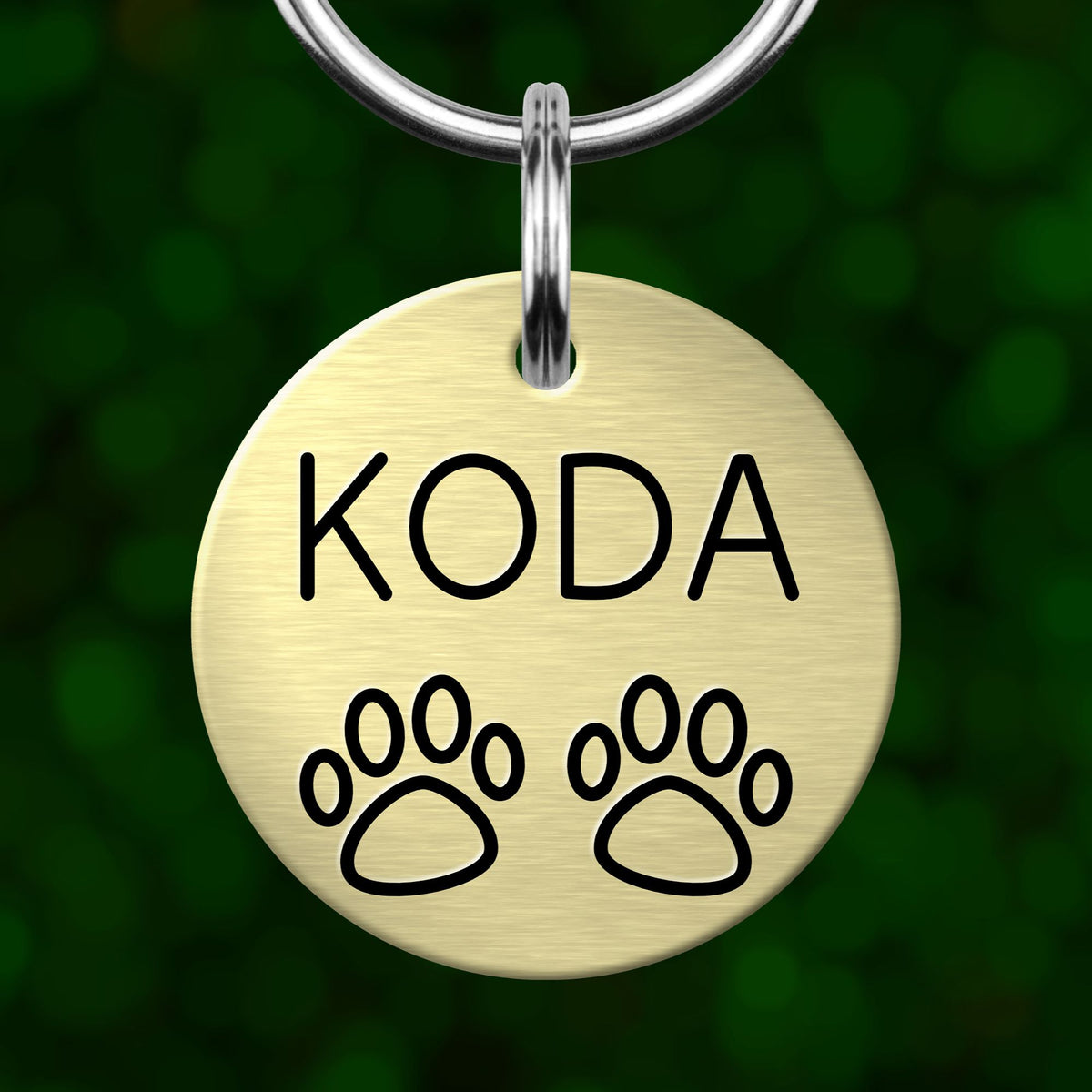 A round metal pet tag with the name "KODA" and paw prints on it, hanging from a keychain.