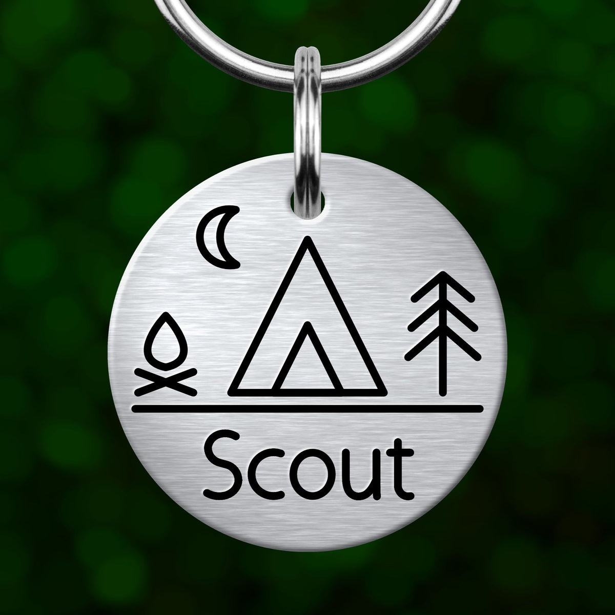 A metal keychain with a circular design featuring a tent, a crescent moon, and a tree, along with the word "Scout" written below.