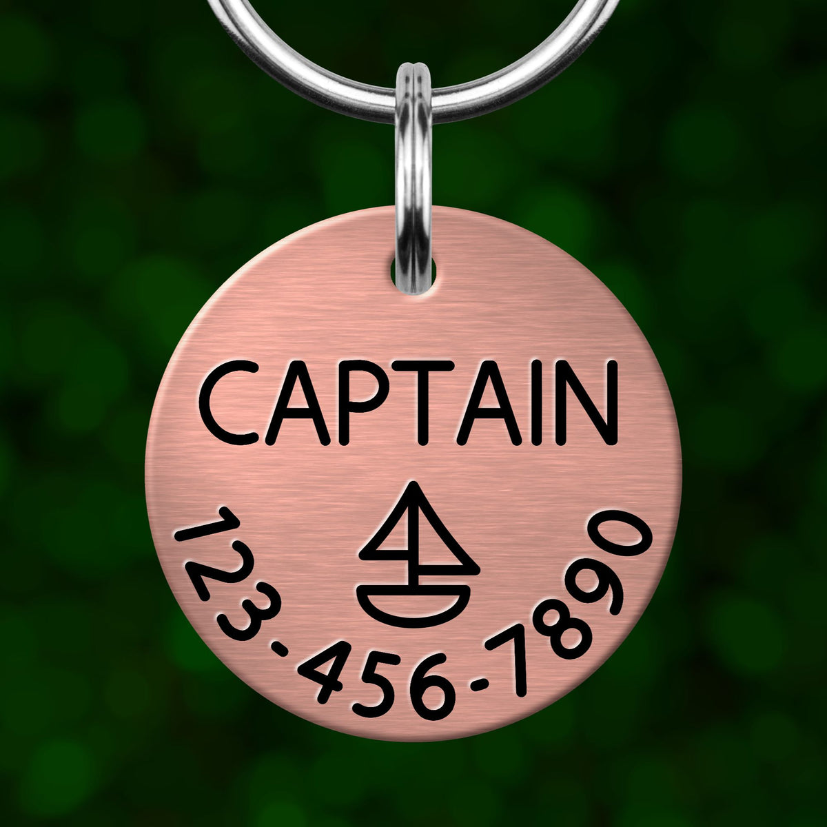 A round metal pet tag with the word "CAPTAIN" printed on it, along with a phone number and a symbol resembling a boat.