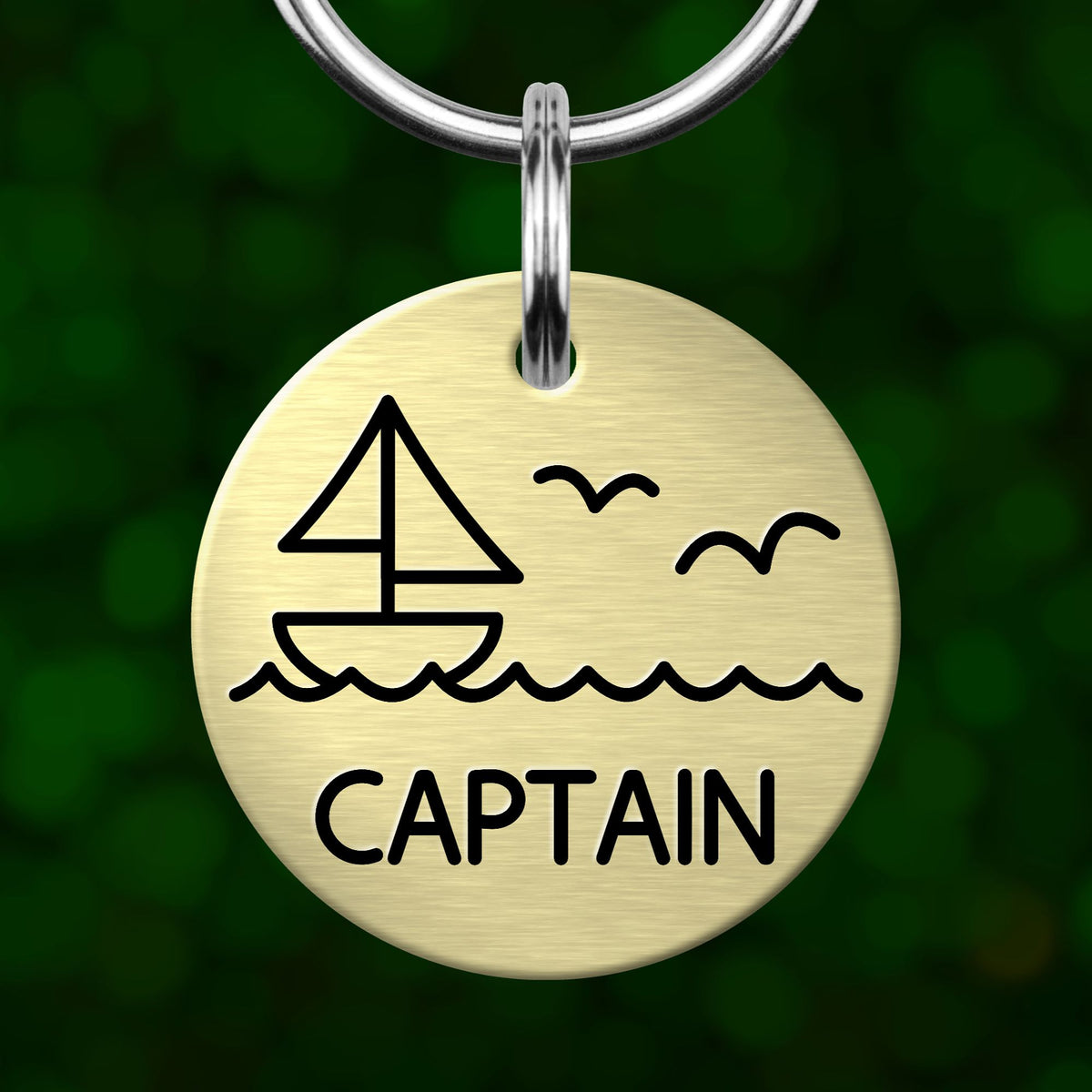 A round metal keychain tag with a graphic design depicting a sailboat and waves, along with the word "CAPTAIN" written below.
