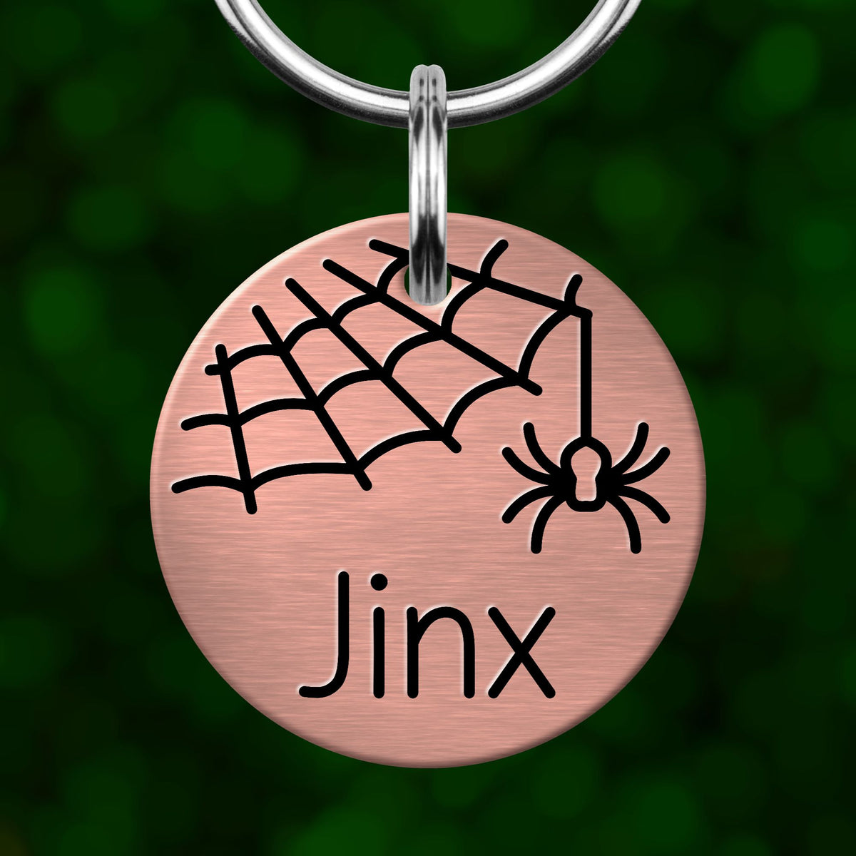 A round metal pet tag with a spider and web design, and the name "Jinx" printed on it.
