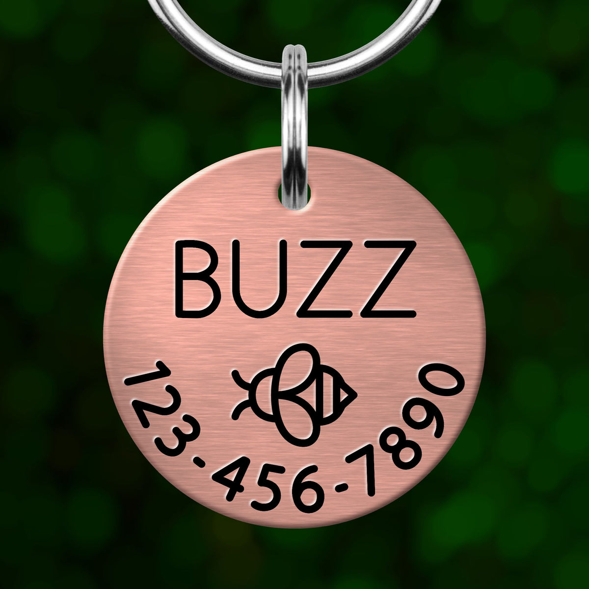A copper-colored pet tag with the name "BUZZ" printed on it, along with a phone number and a bee symbol.