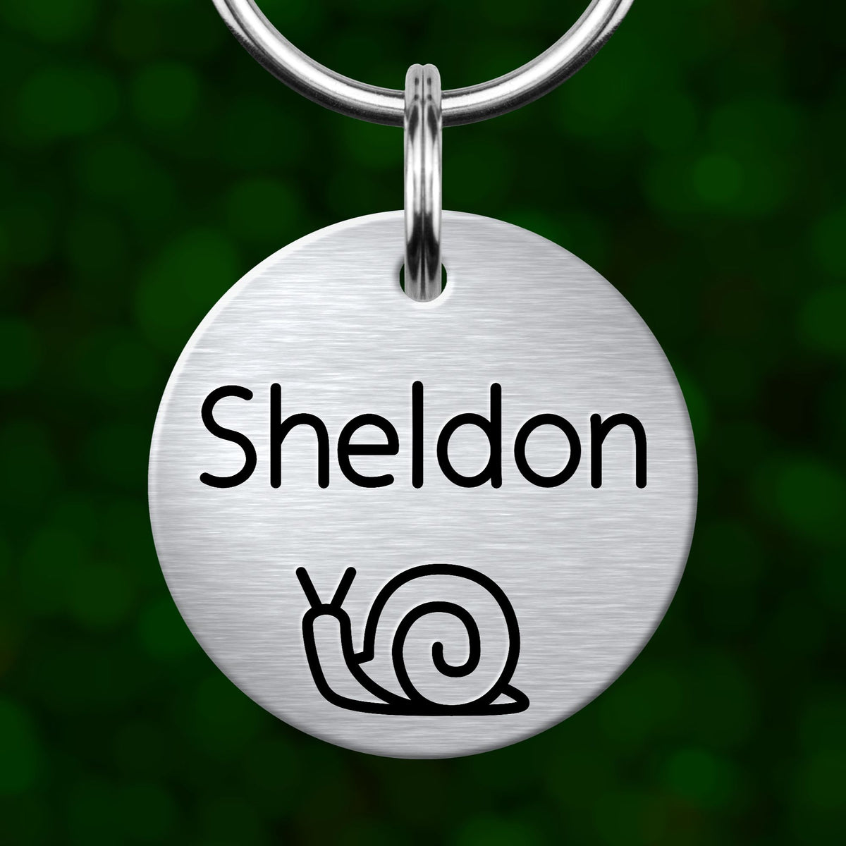 A round metal keychain tag with the name "Sheldon" and a simple black and white illustration of a snail.