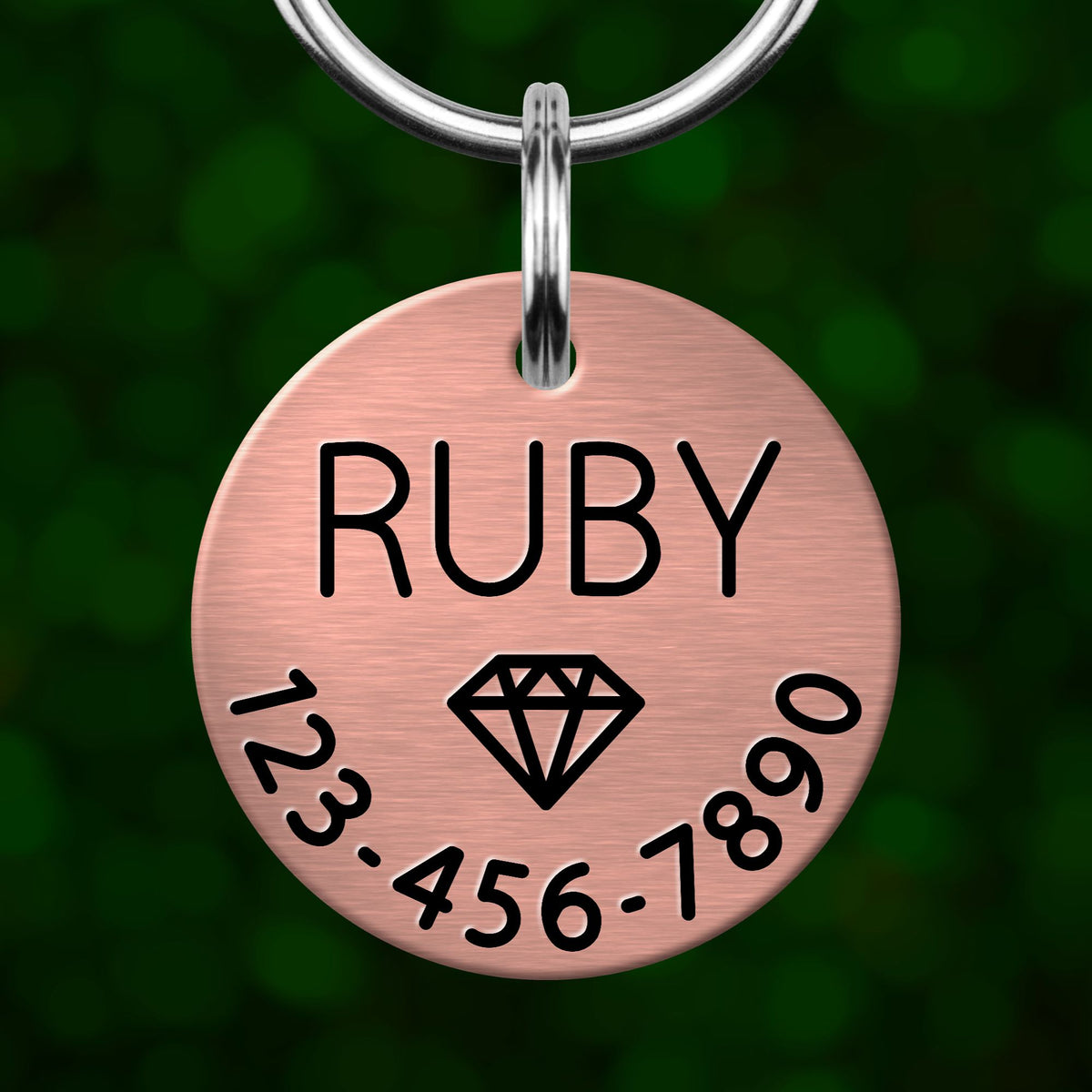 A copper-colored metal tag with the name "Ruby" and a phone number on it, hanging from a keychain.