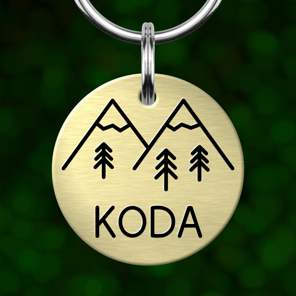A round metal keychain tag with a mountain and tree design, and the word "KODA" written on it.