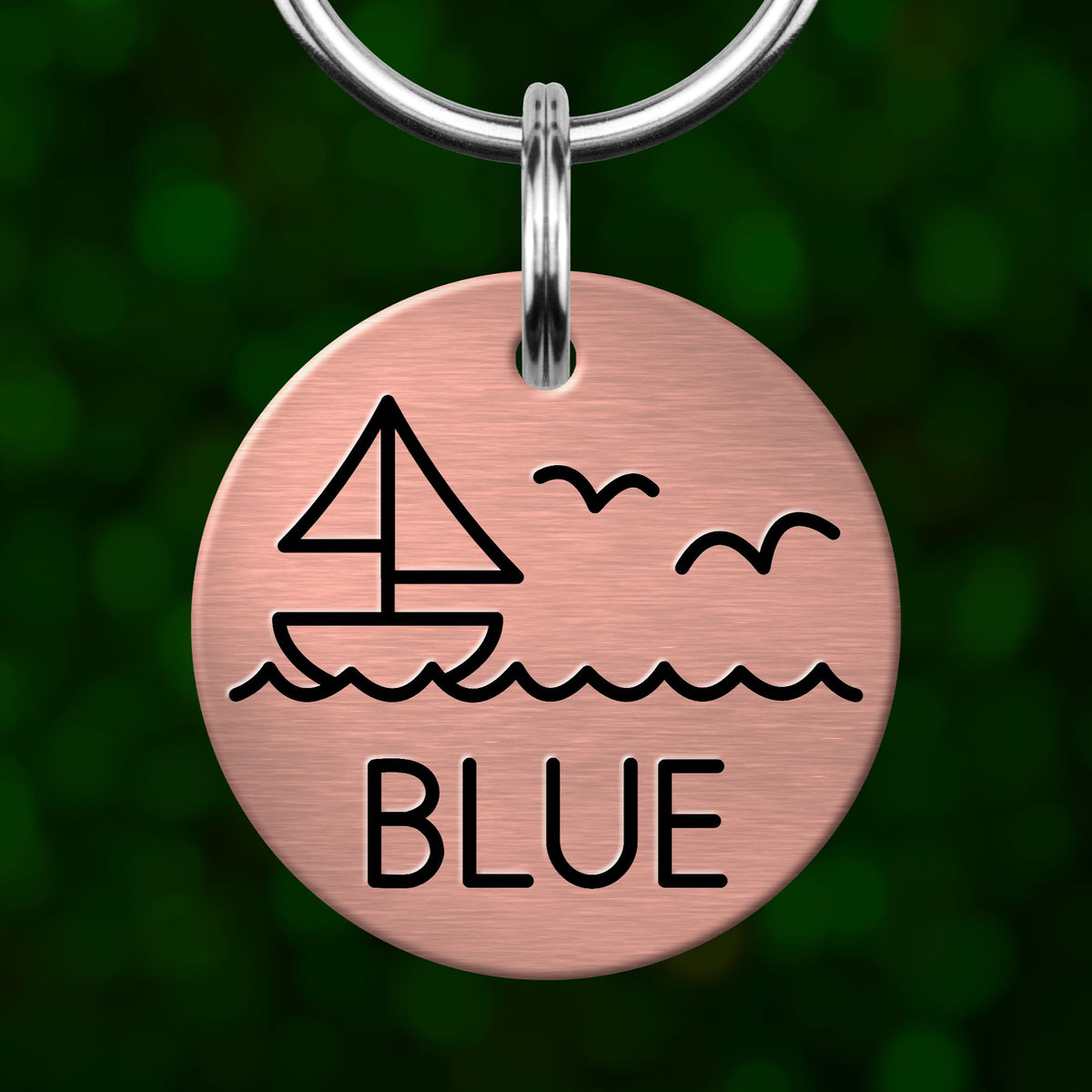 A round metal keychain tag with a design of a sailboat and waves, and the word "BLUE" written on it.