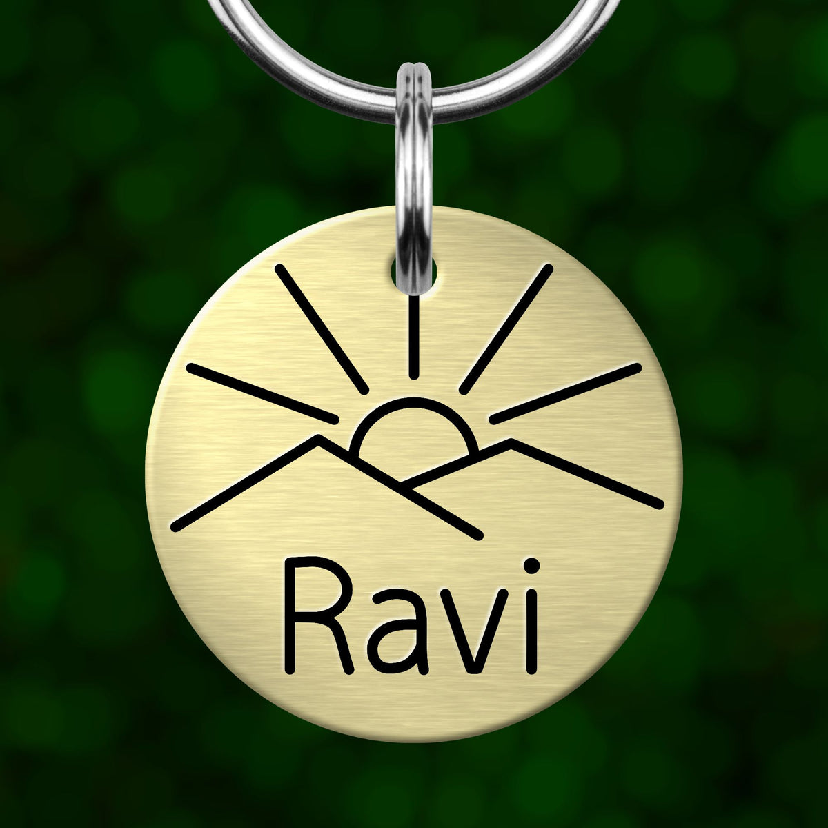 A round metal keychain with a sun and mountain design, and the name "Ravi" written on it.