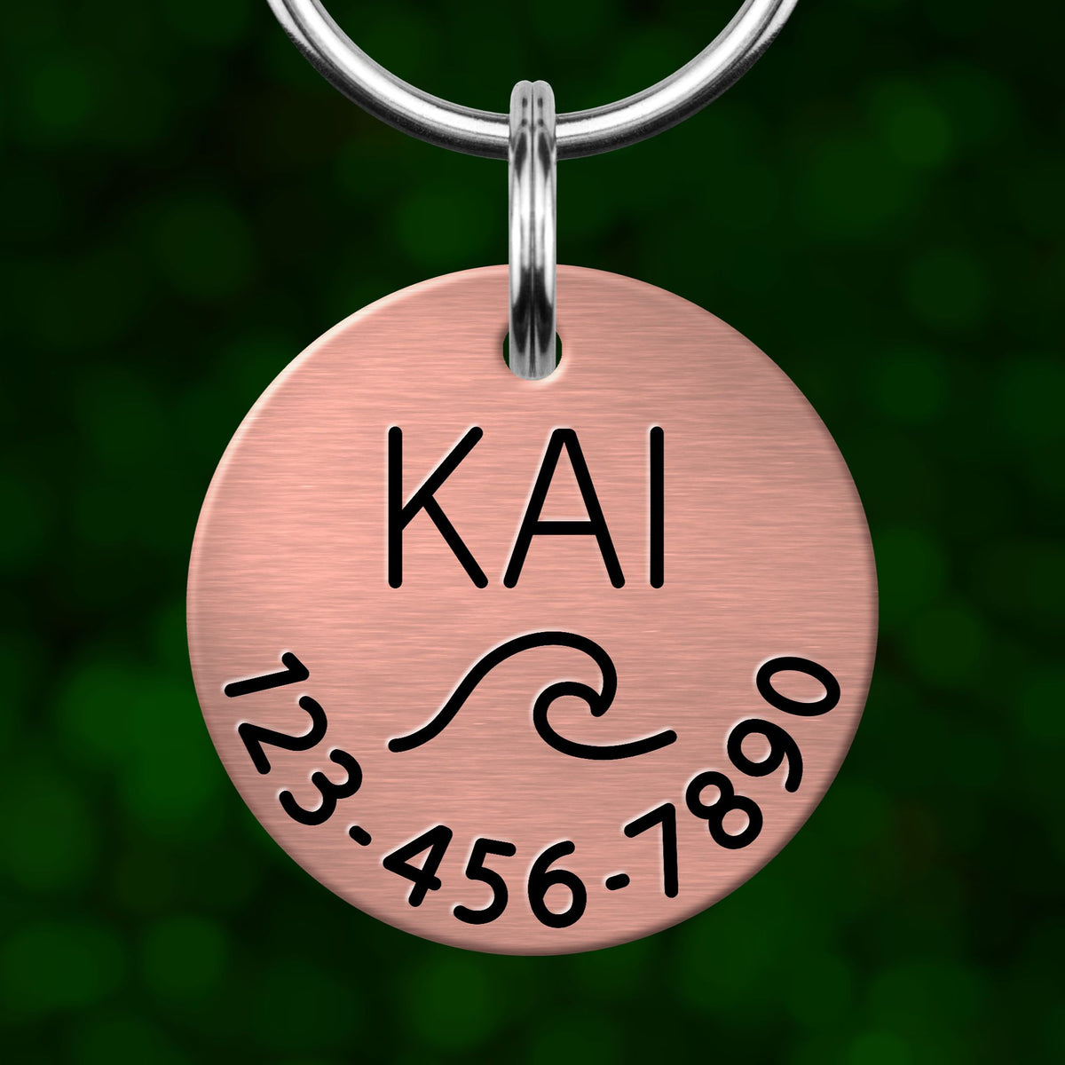 A copper-colored metal pet tag with the name "KAI" and a wave design, along with the numbers "123-456-7890" printed on it.