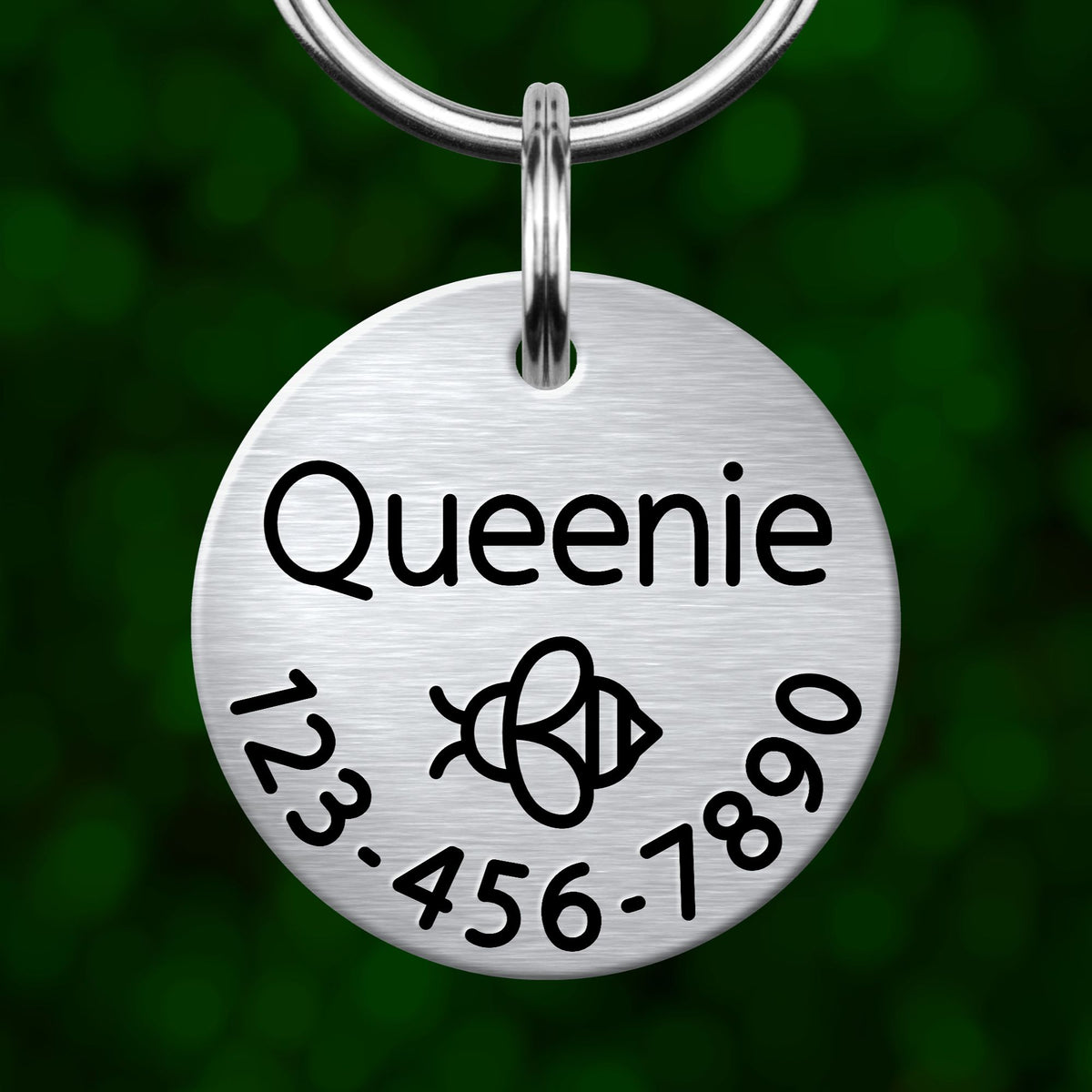 A silver-colored metal tag with the name "Queenie" and a phone number on it, hanging from a keychain.