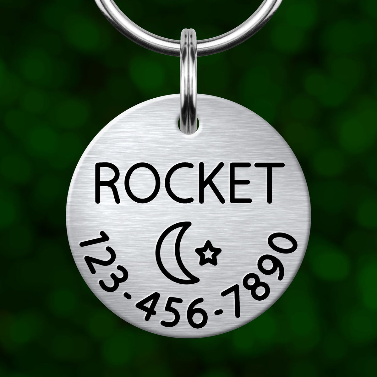 A silver circular pet tag with the word "ROCKET" printed on it, along with a phone number and a symbol of a crescent moon and star.