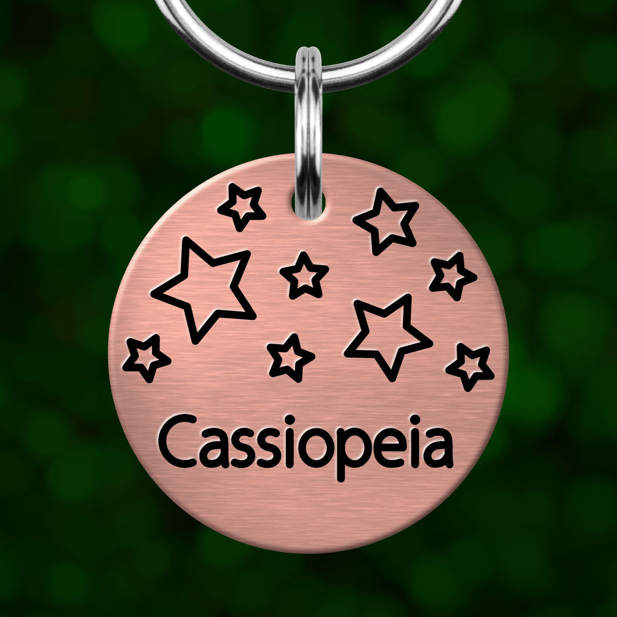 A round metal tag with a metallic appearance, featuring a design of stars and the word "Cassiopeia" written on it.