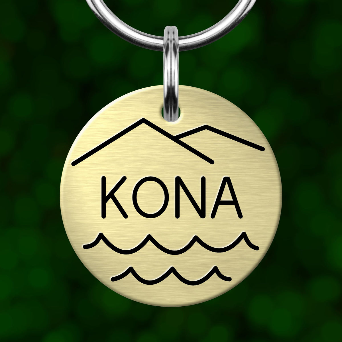 A round metal keychain tag with the word "KONA" printed on it, featuring a mountain and wave design.