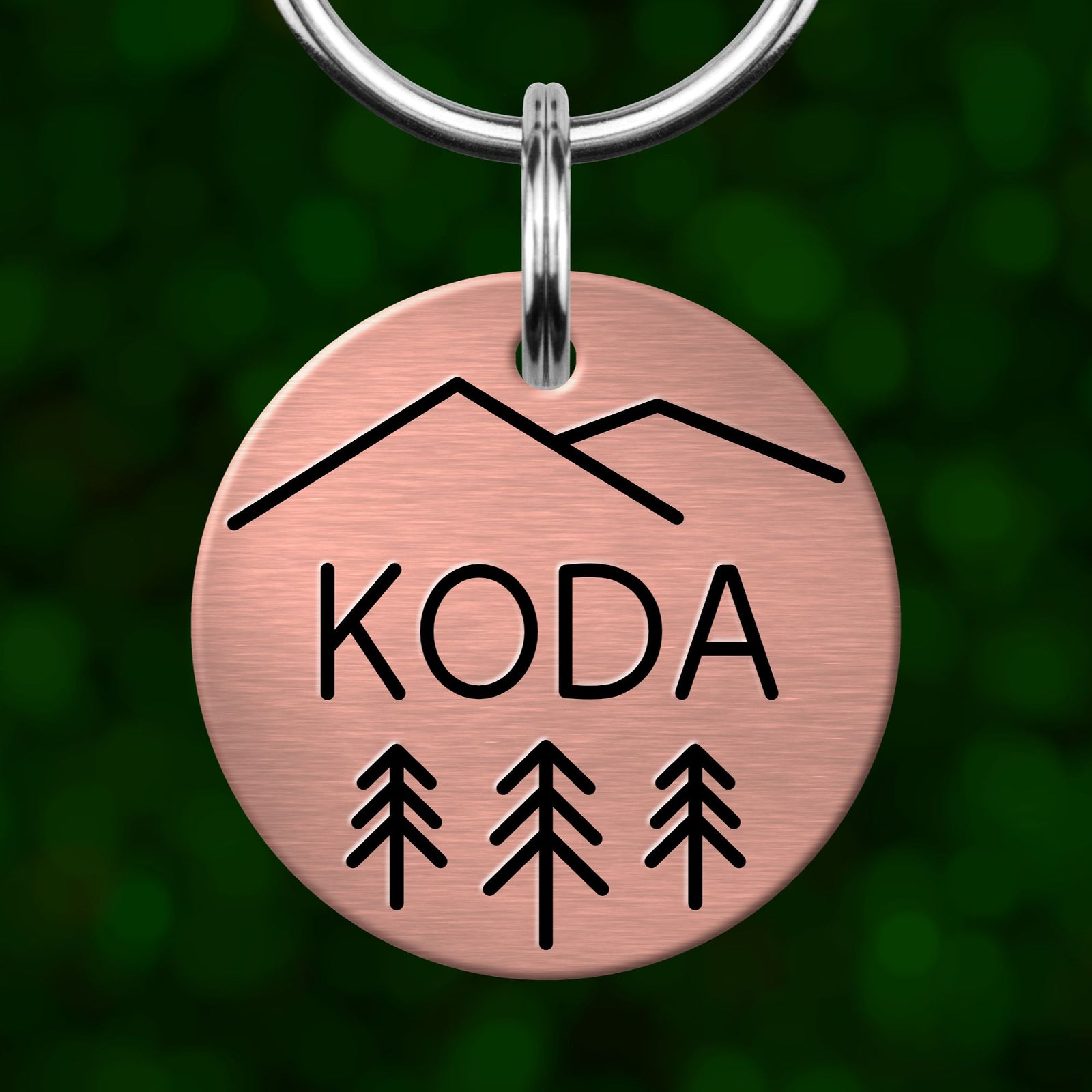 A copper-colored metal keychain with the word "KODA" and three tree silhouettes printed on it.