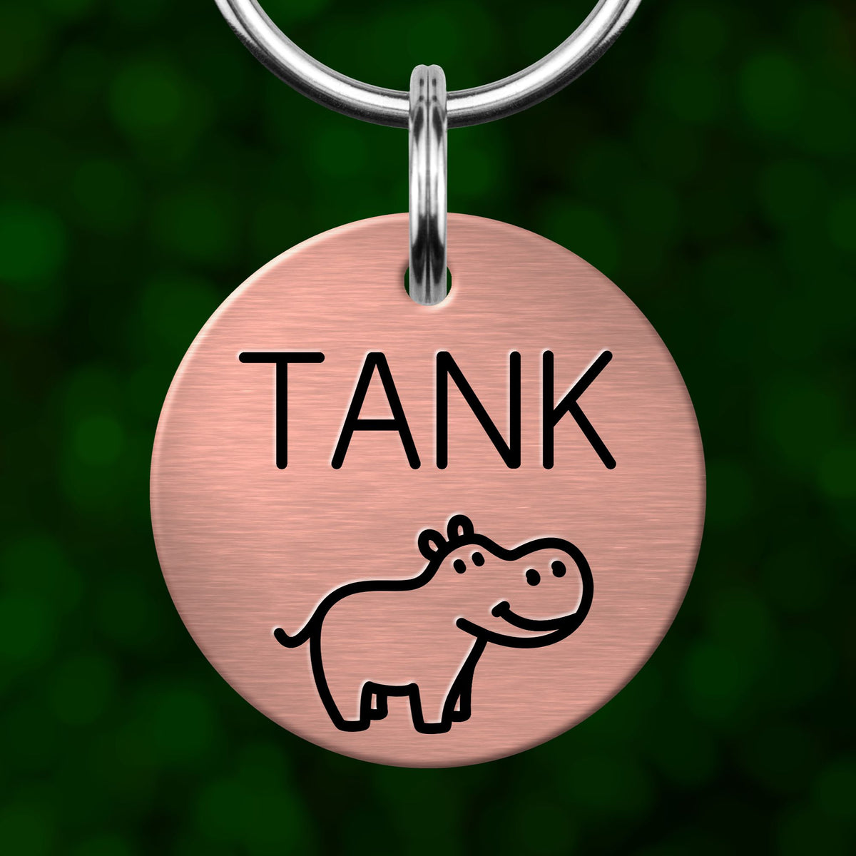 A round metal keychain tag with the word "TANK" printed on it, featuring a small cartoon hippopotamus illustration.