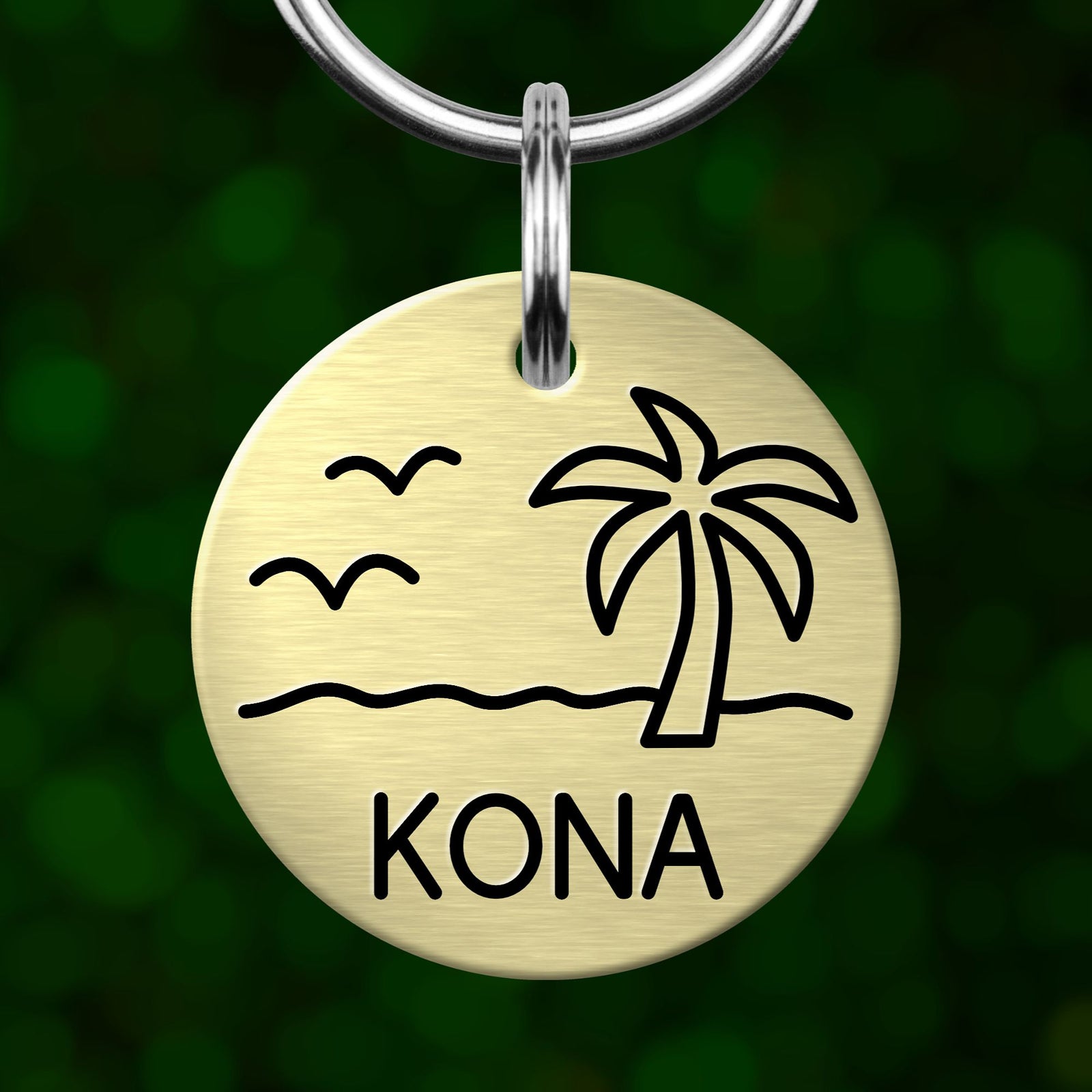 A round metal keychain tag with a beach scene and palm tree design, featuring the word "KONA" below the image.
