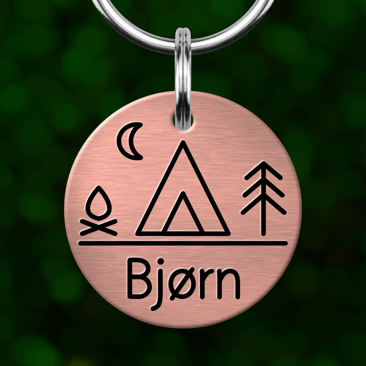 A copper-colored metal keychain tag with a design featuring a camping symbol, a crescent moon, and a tree, along with the name "Bjorn" written below.