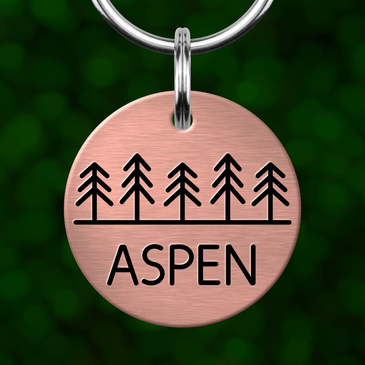 A round metal pet tag with a design of trees and the word "Aspen" printed on it.