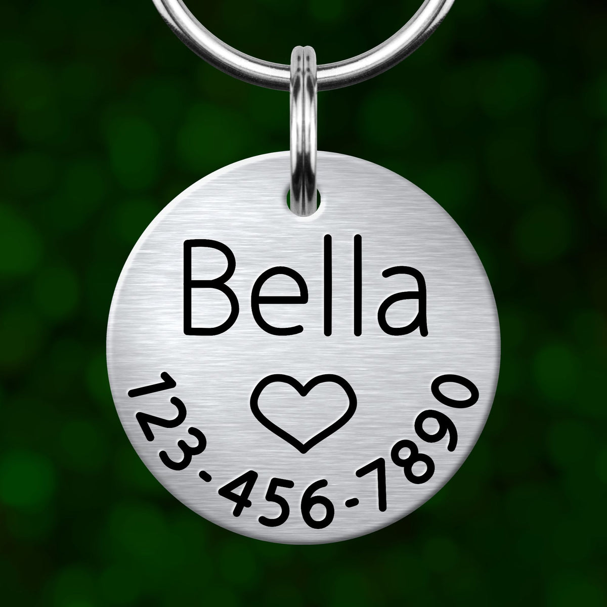A silver-colored metal pet tag with the name "Bella" and a heart symbol, along with a phone number.