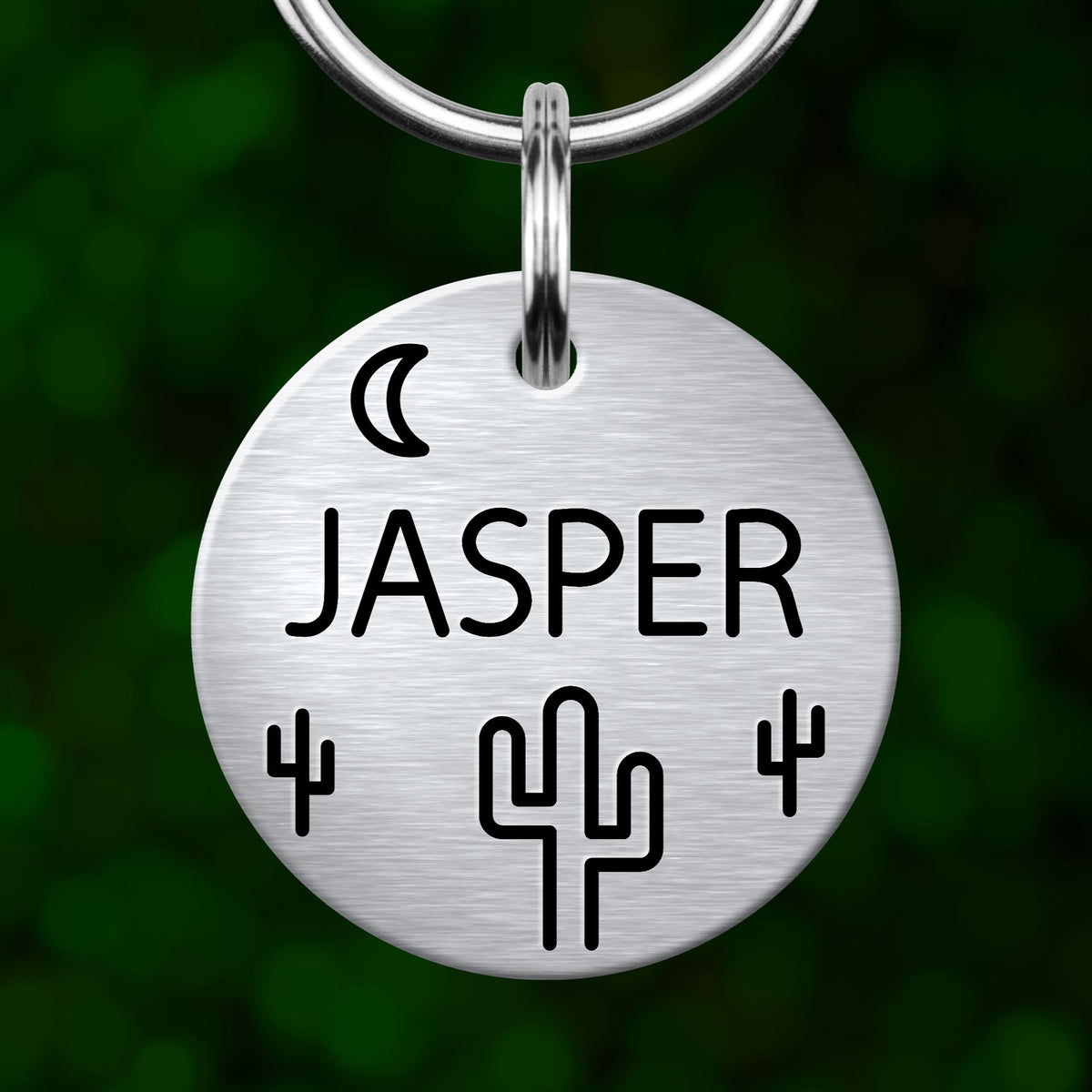 A silver-colored metal name tag with the name "JASPER" printed on it, along with a cactus symbol and a crescent moon.